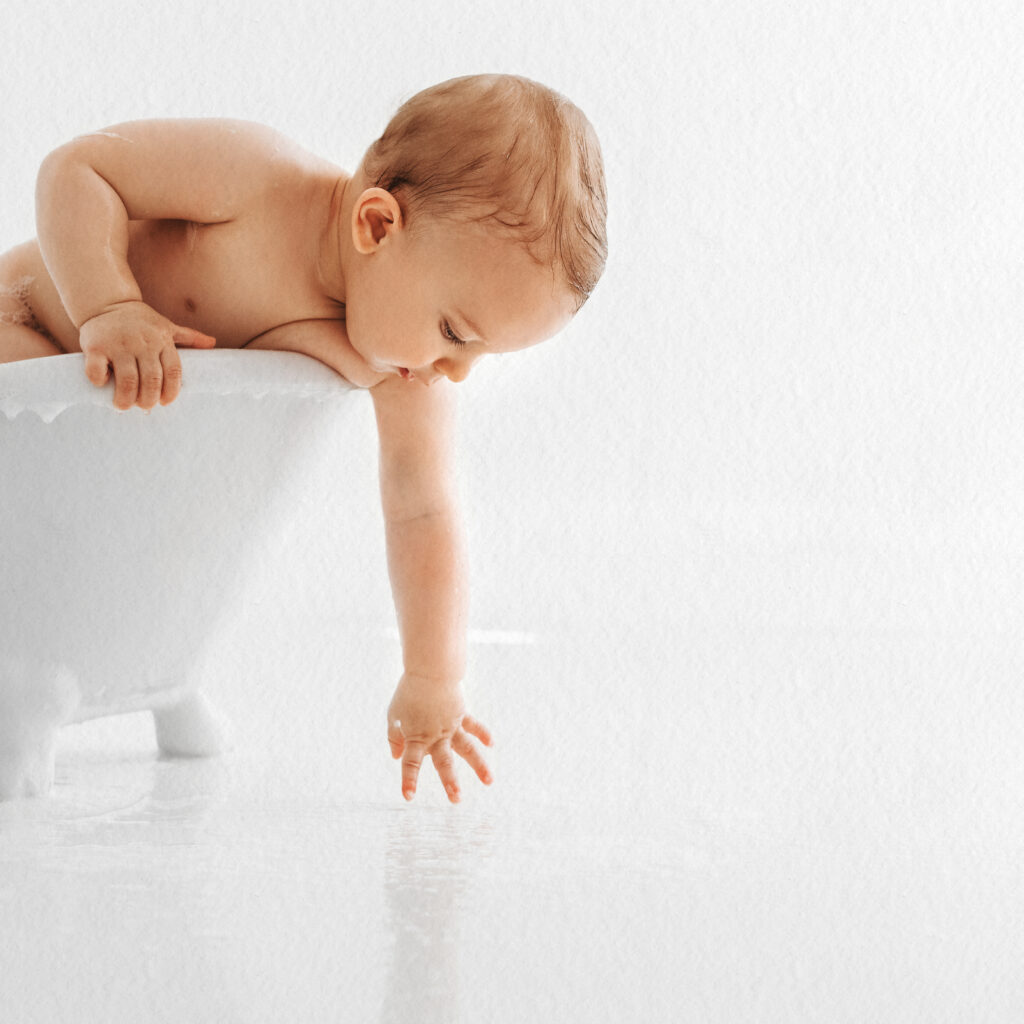 A baby leans over the side of a small white bathtub, reaching down to touch the water on the floor, captured in a playful post-cake smash cleanup moment.