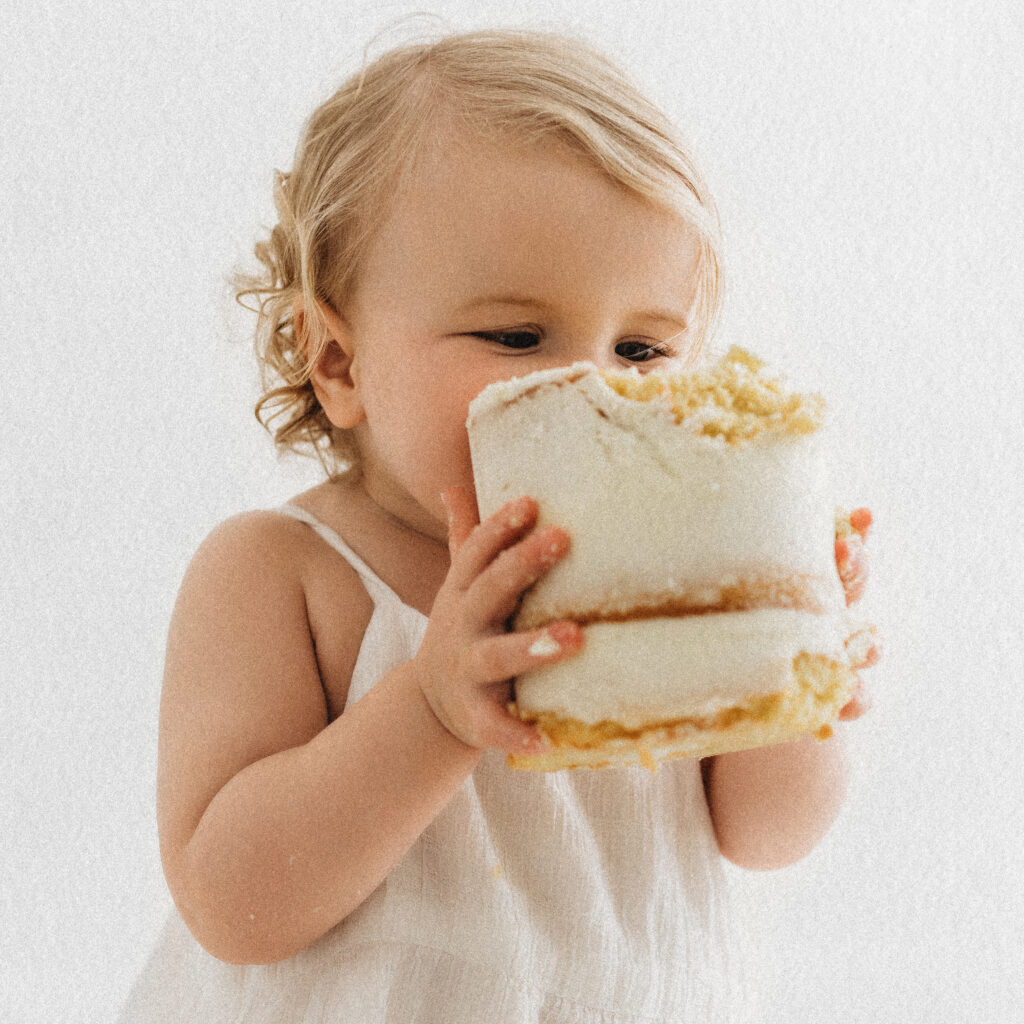 A toddler in a white outfit holds and bites into a small frosted cake, enjoying their first birthday cake smash against a bright white backdrop.