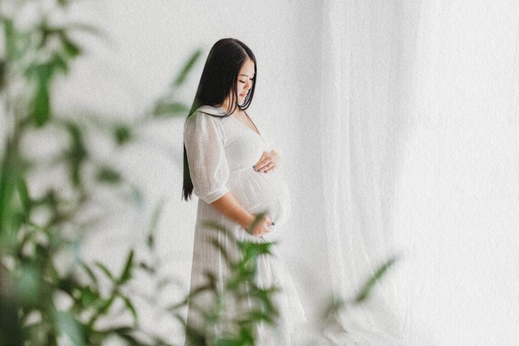 A pregnant woman on a maternity photoshoot  in a flowing white dress cradles her baby bump while standing by soft white curtains, with blurred greenery in the foreground.